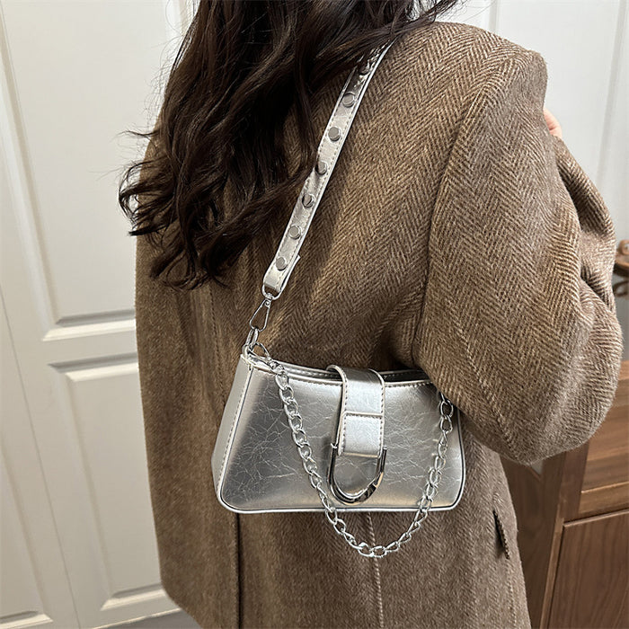 A woman wearing a brown herringbone coat carries a metallic silver shoulder bag with a studded strap and chain detail.