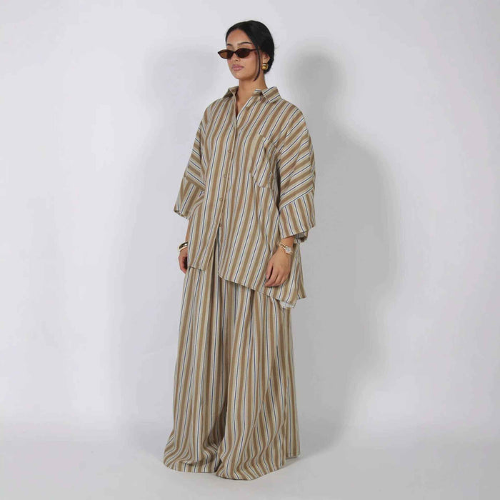 A model wearing a matching beige and brown striped lounge set, featuring a long-sleeve button-up shirt and wide-leg trousers, standing against a plain light gray studio background.