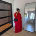 A woman stands in a room wearing a bright red two-piece outfit with a matching red top and wide-leg skirt or pants, paired with a black quilted shoulder bag. She is positioned in front of a large floor mirror and a black-framed glass door.