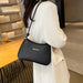 A woman wearing a white coat carries a black crescent-shaped shoulder bag with a small gold bar accent on a warm indoor setting.