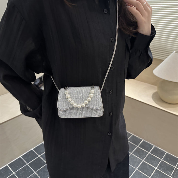 A person wearing a black button-up shirt carries a small silver glittery crossbody bag with a pearl bead detail on the front flap, photographed indoors near a window and lamp.
