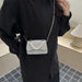 A person wearing a black button-up shirt carries a small silver glittery crossbody bag with a pearl bead detail on the front flap, photographed indoors near a window and lamp.