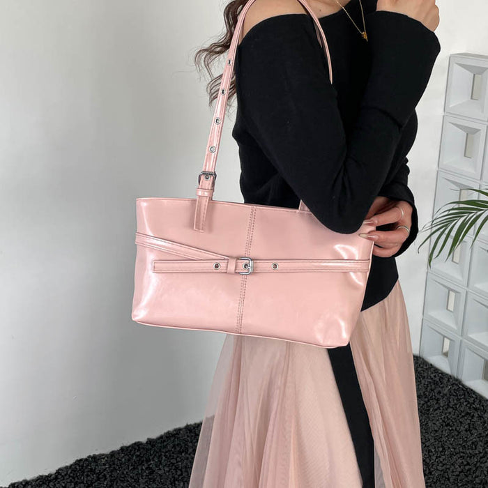 A woman wearing a black off-the-shoulder top and a sheer light pink skirt carries a glossy light pink shoulder bag with silver-tone hardware in an indoor setting.