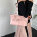 A woman wearing a black off-the-shoulder top and a sheer light pink skirt carries a glossy light pink shoulder bag with silver-tone hardware in an indoor setting.