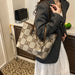 A woman wearing a gray blazer and white skirt carries a large beige tote bag with a brown monogram pattern and brown leather straps in an indoor setting.