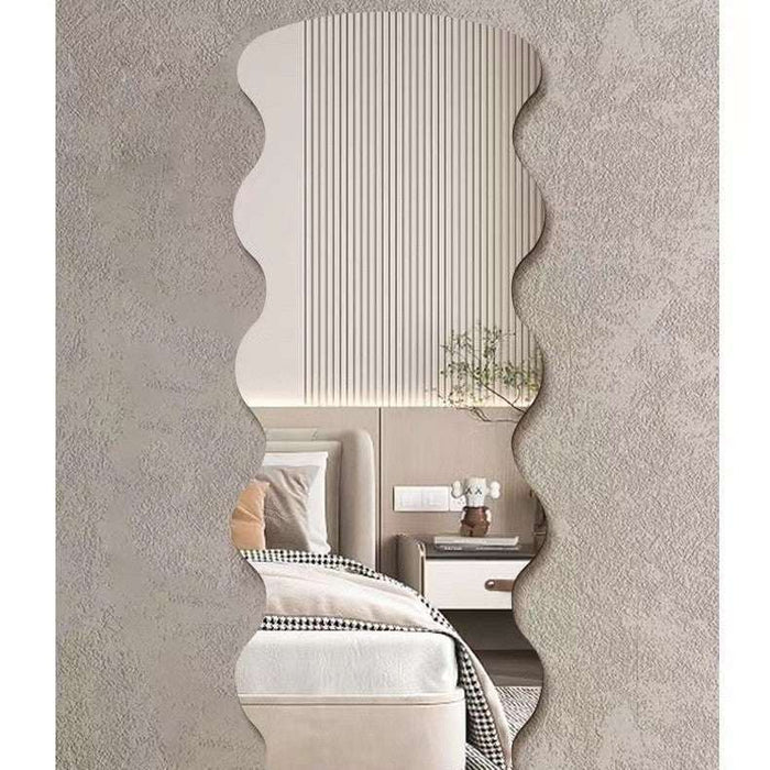 A wavy, scalloped-edge wall mirror with a textured beige frame, shown mounted on a textured gray wall and reflecting a modern bedroom interior.