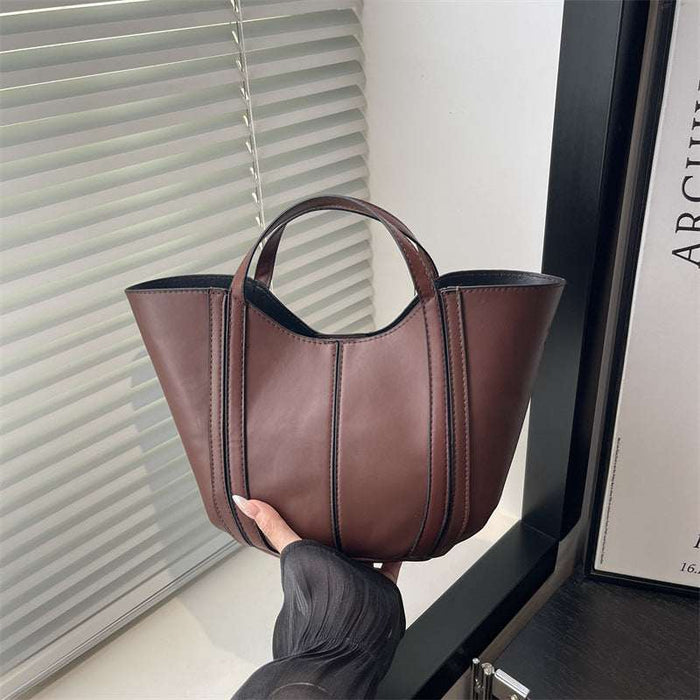 A brown leather tote bag with double top handles, held by a hand in front of a window with white blinds.