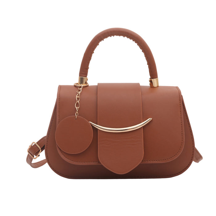 A brown faux-leather handbag with a structured top handle, a curved gold-tone metal accent on the front flap, a matching round hanging charm, and a shoulder strap, shown on a plain white background.