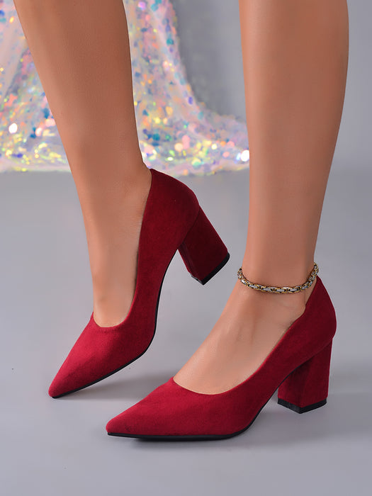 A pair of red suede block-heel pumps with pointed toes, shown on a model's feet against a gray studio background with a sparkly sequined fabric backdrop.