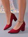 A pair of red suede block-heel pumps with pointed toes, shown on a model's feet against a gray studio background with a sparkly sequined fabric backdrop.
