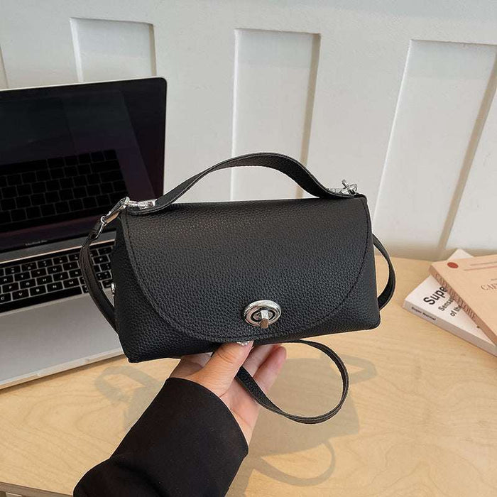 A black pebbled faux-leather handbag with a top handle and silver-tone hardware, held by a hand over a light wood desk with a laptop and books in the background.