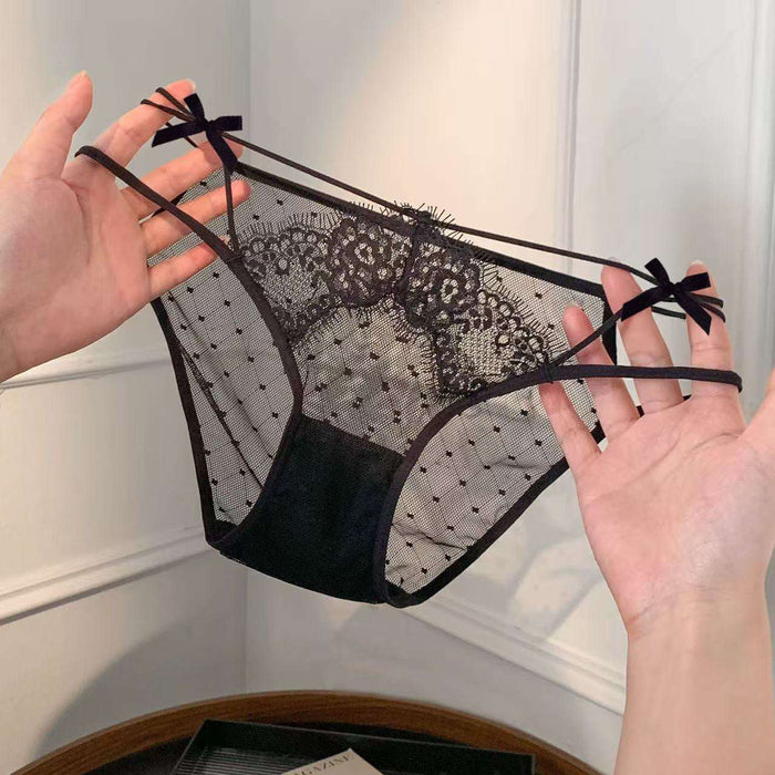 Black lace and mesh panties with thin straps and small bow details, held up by two hands against an indoor wall background.