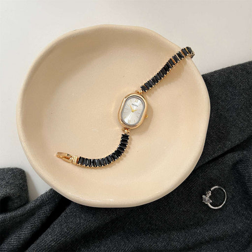 Gold watch with black beaded strap on a beige dish, gray fabric background