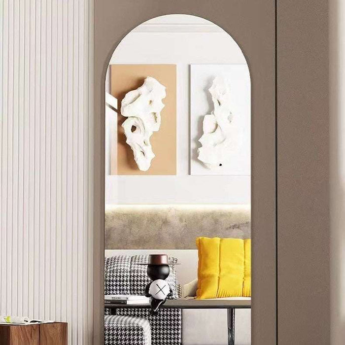 An arched wall mirror with a slim frame hangs on a taupe wall, reflecting a modern living room interior with abstract wall art, a sofa, and a yellow pillow.