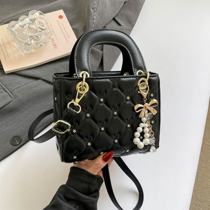 A black quilted top-handle handbag with gold-tone hardware, star details, and a pearl bow charm, held by a hand against a styled background of a gray blazer and white surface.