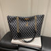 A black and gray monogram pattern tote bag with gold chain straps and a deer charm, displayed on a white book resting on a black leather seat.