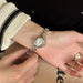 A silver-toned watch with a round face and decorative links is worn on a person's wrist, with hands adjusting it. The background includes a cream knit sweater, black clothing, and a beige surface.