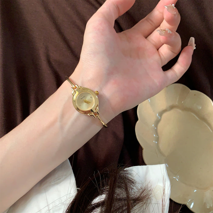 A gold-toned watch with a round dial and decorative side details is worn on a person's wrist, shown against a dark brown fabric background with a cream dish nearby.