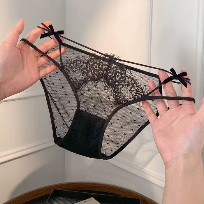Black lace and mesh panties with thin straps and small bow details, held up by two hands against an indoor wall background.