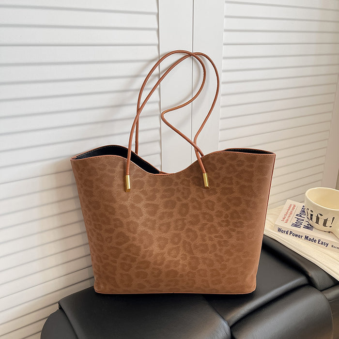 A brown leopard-print tote bag with double shoulder straps and gold-tone hardware, resting on a black leather chair in front of white slatted doors.