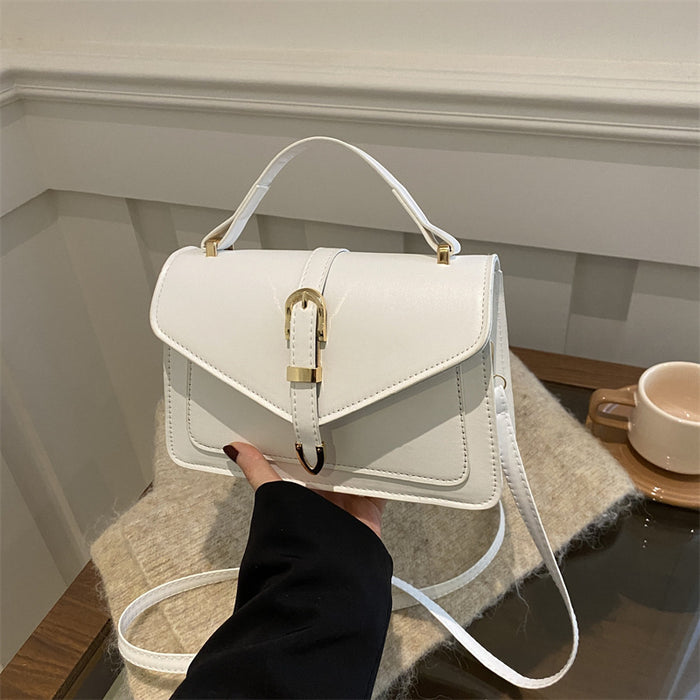 A white faux leather handbag with a top handle and a gold buckle closure, held by a hand wearing a black sleeve. The bag is resting on a textured beige blanket on a glass table with a coffee cup in the background.