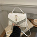 A white faux leather handbag with a top handle and a gold buckle closure, held by a hand wearing a black sleeve. The bag is resting on a textured beige blanket on a glass table with a coffee cup in the background.