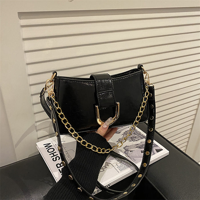 A black faux-leather shoulder bag with a gold-tone chain strap and gold studs, held by a hand over a magazine on a black leather surface.
