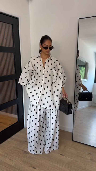 A woman stands indoors wearing a white matching lounge set with black polka dots, consisting of a long-sleeve button-front top and wide-leg pants. She is holding a small black handbag and wearing dark sunglasses.