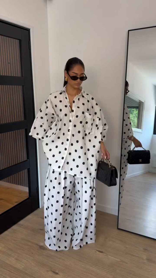 A woman stands indoors wearing a white matching lounge set with black polka dots, consisting of a long-sleeve button-front top and wide-leg pants. She is holding a small black handbag and wearing dark sunglasses.