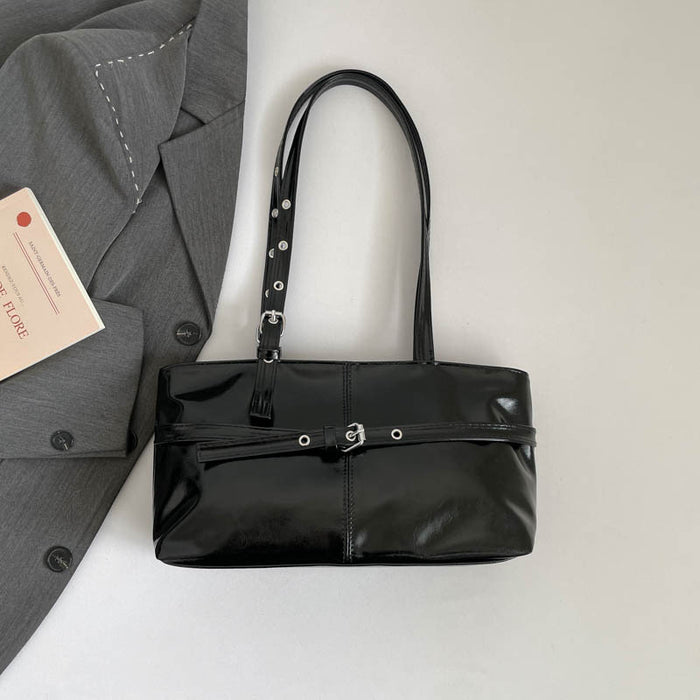 A glossy black patent leather shoulder bag with silver-tone hardware, laid flat on a light gray surface next to a gray blazer and a small card.
