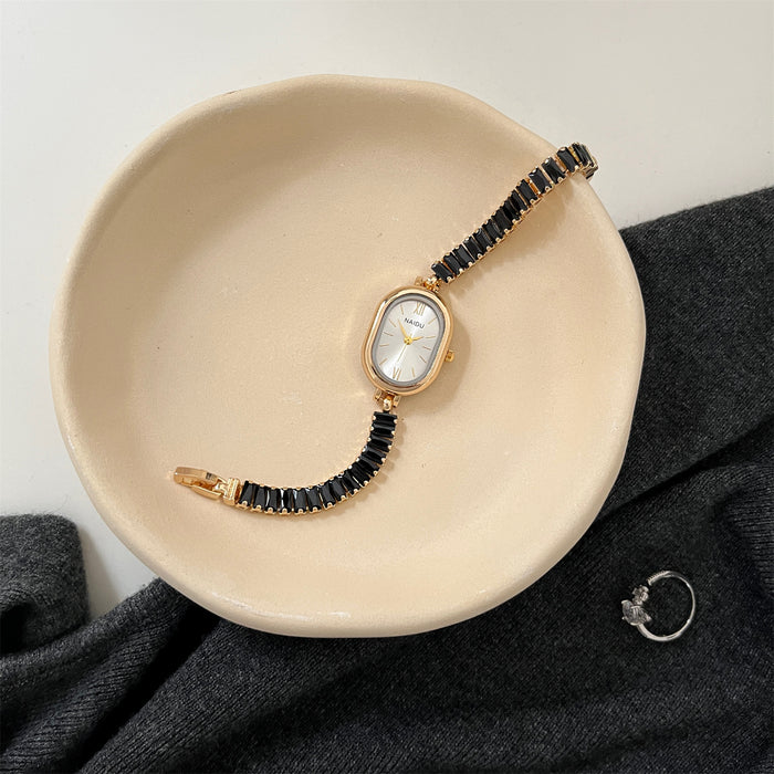 Gold watch with black beaded strap on a beige dish, gray fabric background