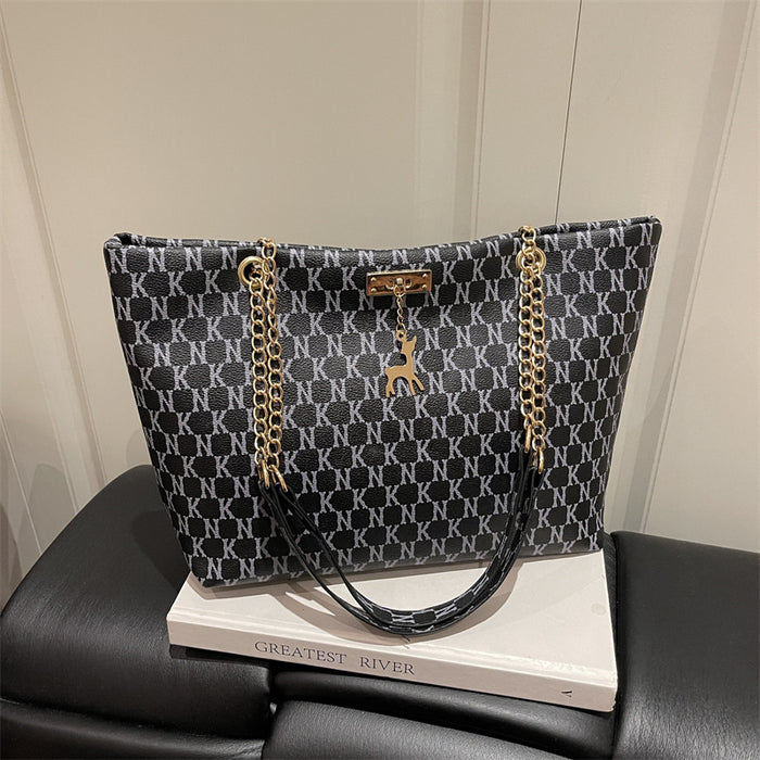 A black and gray monogram pattern tote bag with gold chain straps and a deer charm, displayed on a white book resting on a black leather seat.