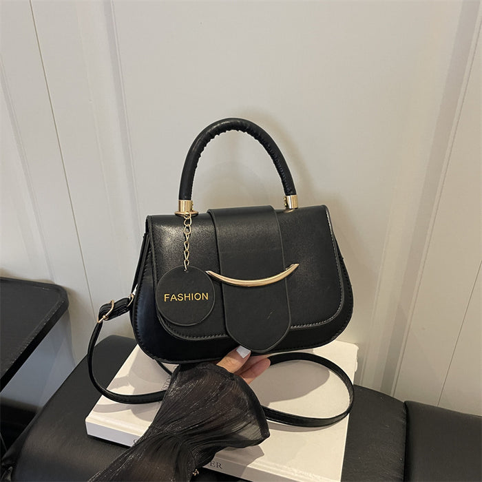 A black leather handbag with a top handle, gold-tone hardware, and a hanging circular charm that reads 'FASHION', shown resting on a white box with a hand holding the strap in a modern indoor setting.