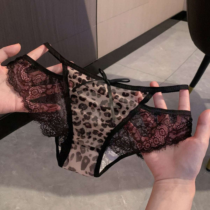 A pair of sheer leopard-print lace panties with black trim and floral lace side panels, held up by two hands against an indoor tiled floor background.
