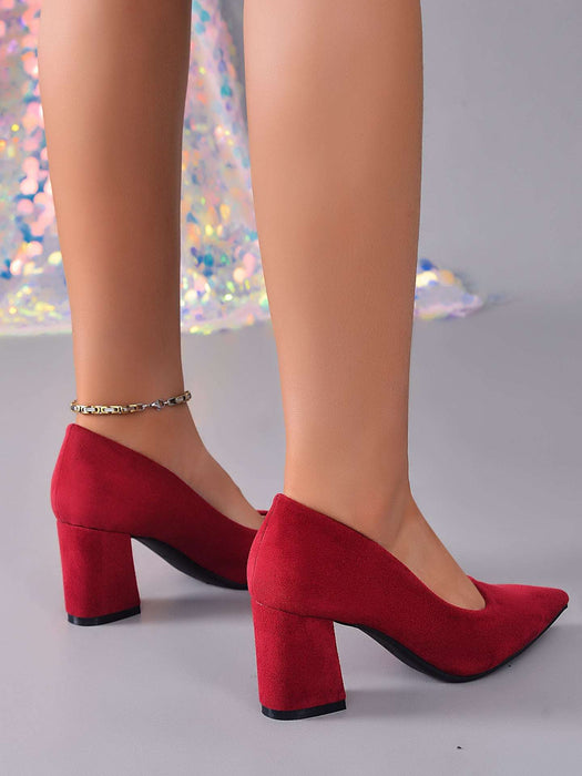 A pair of red suede block-heel pumps with pointed toes, shown on a mannequin or model's feet against a light gray background with a sparkly decorative element.