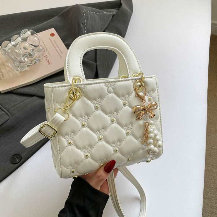 A white quilted top-handle handbag with gold-tone hardware, pearl embellishments, and a dangling bow-and-pearl charm, held by a hand with red nail polish against a white tabletop with a gray blazer and glass bottle in the background.