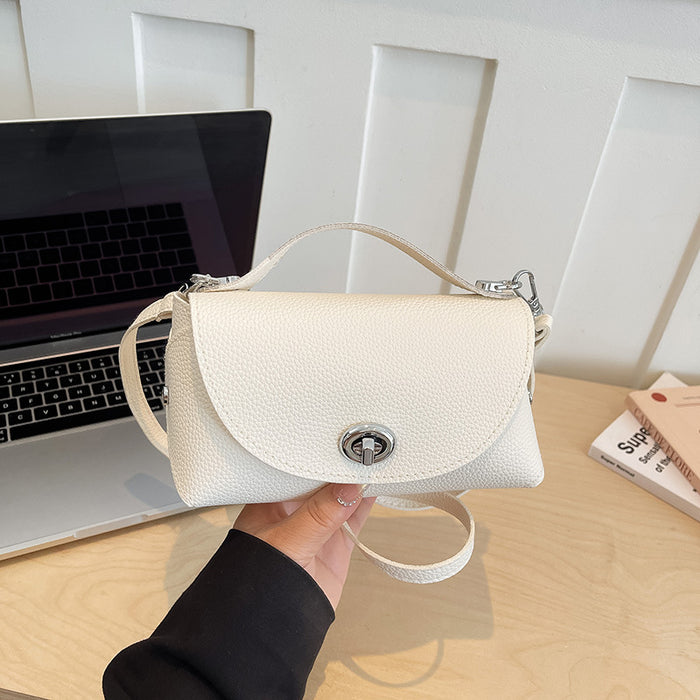 A hand holding a small off-white pebbled leather handbag with a top handle and silver hardware, set against a desk background with a laptop and books.