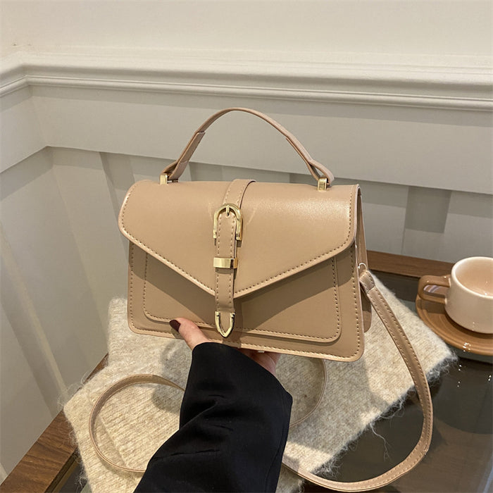 A beige faux leather handbag with a top handle, flap closure, and gold buckle detail, held by a person wearing a black sleeve in an indoor setting.