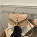 A beige faux leather handbag with a top handle, flap closure, and gold buckle detail, held by a person wearing a black sleeve in an indoor setting.