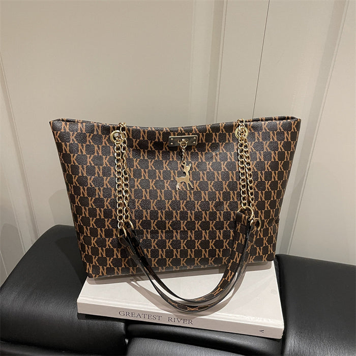 A brown and black monogram patterned tote bag with gold-tone chain straps and a small deer-shaped charm, displayed on a white book resting on a black leather sofa.