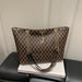 A brown and black monogram patterned tote bag with gold-tone chain straps and a small deer-shaped charm, displayed on a white book resting on a black leather sofa.