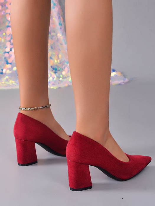 A pair of red suede block-heel pumps with pointed toes, shown on a mannequin or model's feet against a light gray background with a sparkly decorative element.