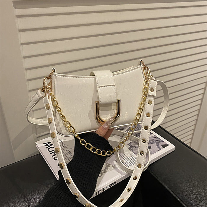 A white faux leather shoulder bag with a gold-tone metal D-ring clasp and a gold chain strap, styled on a black leather surface with a magazine in the background.