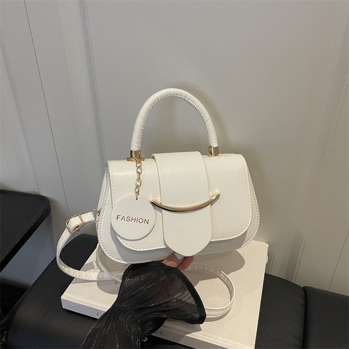 A white faux-leather handbag with a top handle, gold-tone hardware, and a round hanging charm that reads 'FASHION', shown resting on a white box with a black skirt prop in front.