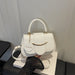 A white faux-leather handbag with a top handle, gold-tone hardware, and a round hanging charm that reads 'FASHION', shown resting on a white box with a black skirt prop in front.