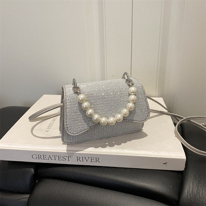 A small light gray woven handbag with a pearl bead strap, resting on a white book with the title 'GREATEST RIVER' on a black leather chair.