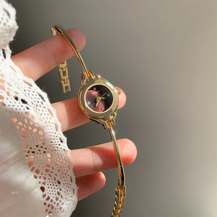 A gold-tone bracelet watch with a round floral dial, held in a hand wearing a white lace sleeve against a soft gray background.