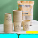 Bamboo fiber paper cup set with a large printed packaging bag and multiple stacks of disposable cups on a white surface against a green wall. Chinese text appears on the bag and as an overlay banner.