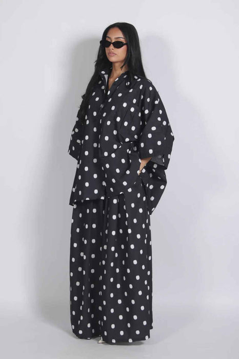 A woman modeling a black and white polka dot matching lounge set consisting of a long-sleeve button-up shirt and wide-leg pants, styled with black sunglasses against a light gray studio background.