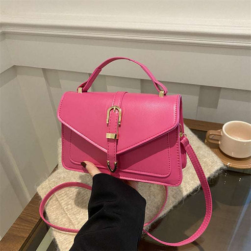A bright pink structured handbag with a top handle, flap front, and gold-tone clasp, held by a hand over a cream knit blanket in an indoor setting.